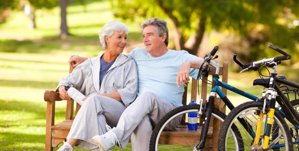 two active seniors sitting on a park bench next to their mountain bikes